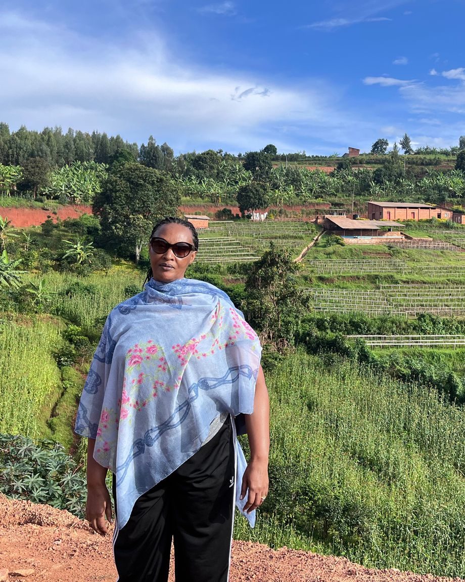 Immy Numutako standing above terraced highland farmland in Rwanda, with coffee drying beds and farm buildings on the slope behind her.